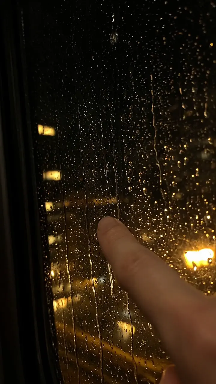 Rain on a dark window with golden city lights behind it and a fingertip touching the glass.